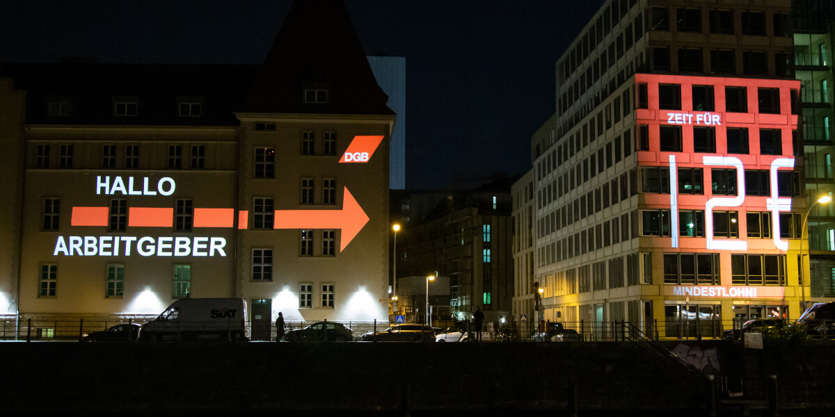 Lichtaktion: Leuchtschrift "Hallo Arbeitgeber, Zeit für 12 Euro Mindestlohn" an Hausfassade