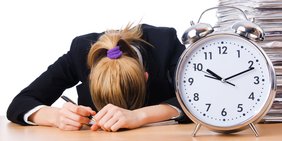 Frau liegt mit Kopf (schlafend) auf Schreibtisch; daneben eine übergroßer Wecker