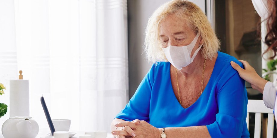 ältere Frau sitzt alleine am Tisch mit Nasen- und Mundschutz