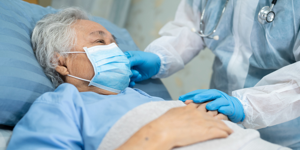 Alte Frau mit Mund-Nasen-Schutz in Krankenhaus-Bett; Hände von Arzt/Ärztin mit Kittel und Stetoskop
