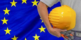 Nahaufnahme eines Schutzhelms, den ein Bauarbeiter auf dem Hintergrund der EU-flagge hält