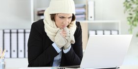 Junge Frau sitzt mit Mütze, Schal und Handschuhen vor ihrem Laptop in einem Büro.