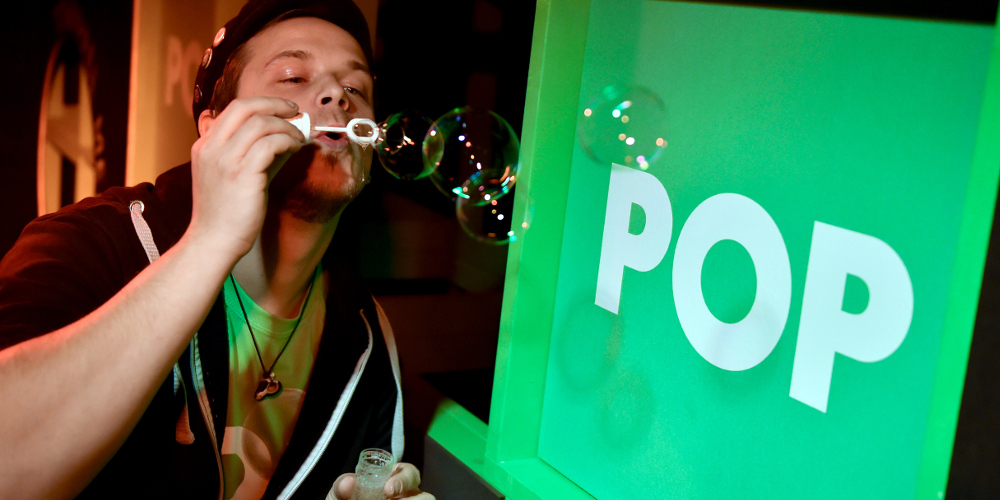Mann bläst Seifenblasen in Richtung eines grünen Würfels mit der weißen Aufschrift "POP"