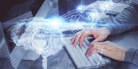 Hände arbeiten am Laptop und darüber liegt ein Hologramm von einem meschanischem Arm und einem Gehirn