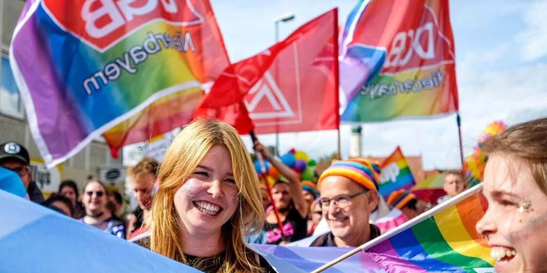 Eine junge lachende Frau mit Glitzer im Gesicht und Regenbogenflagge auf CSD in Landshut, im Hintergrund Regenbogenflagge mit Aufschrift "DGB Niederbayern" und rote Flagge mit dem Logo der IG Metall
