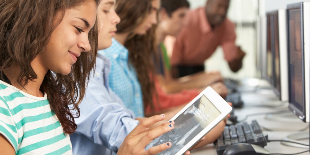 Im Vordergrund: Lächelndes Mädchen mit Tablet; im Hintergrund: Weitere Schülerinnen und Schüler sowie ein Lehrer an Computer-Bildschirmen