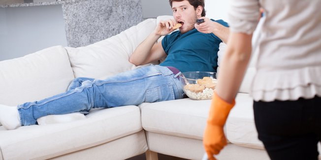 Mann liegt auf Sofa, schaut Fernsehen und isst Chips, Frau arbeitet im Haushalt