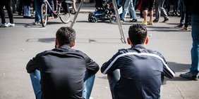Zwei Männer in öhnlichem Outfit sitzen auf der Stufe in einer Fußgängerzone, Rückenansicht