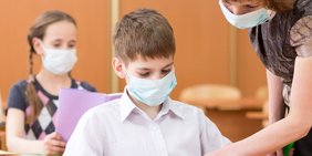 Kinder mit Mund-Nasenschutz in der Schule mit Lehrerin