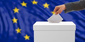 Mann steckt Umschlag in eine Wahlurne, die vor einer Europa-Flagge steht