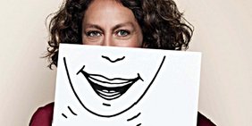 Frau hält Blatt Papier mit gezeichnetem lachenden Mund vor ihren eigenen Mund