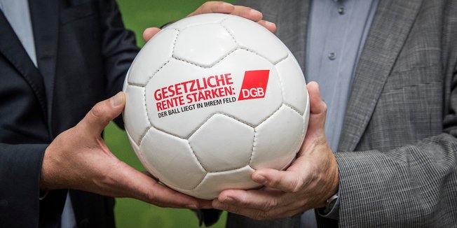 Vier Hände halten einen weißen Lederfußball mit der Aufschrift "Gesetzliche Rente stärken: Der Ball liegt in Ihrem Feld"; daneben das DGB-Logo