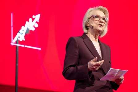 Monika Schnitzer mit blonden Haaren und Brille hält Unterlagen in der Hand, hinter ihr ein roter Hintergrund mit Wachstumspfeil-Symbol.