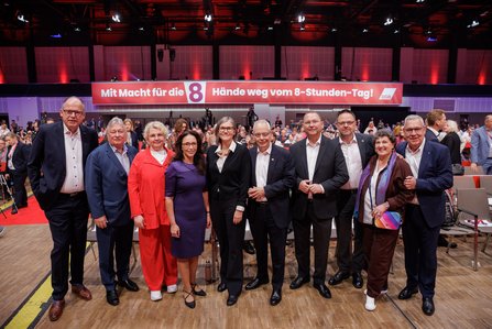 Der DGB-Bundesvorstand steht in einer Reihe vor dem gefüllten Kongresssaal, v.l.n.r.: Stefan Körzell (DGB), Martin Burkert (EVG), Maike Finnern (GEW), Yasmin Fahimi (DGB-Vorsitzende), Christiane Benner (IG Metall), Michael Vassiliadis (IGBCE), Frank Werneke (ver.di), Guglielmo Mangiapane (NGG), Anja Piel (DGB), Robert Feiger (IG BAU); im Hintergrund ein Banner mit dem Slogan "Mit Macht für die 8 – Hände weg vom 8-Stunden-Tag!".
