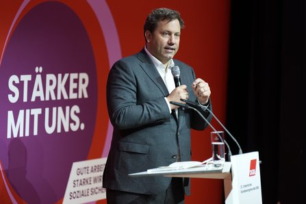 Ein Mann in grauem Anzug und weißem Hemd steht mit einem Mikrofon in der Hand und gestikuliert. Hinter ihm ist eine Leinwand mit dem Schriftzug „STÄRKER MIT UNS." zu sehen. Vor ihm steht ein Rednerpult.