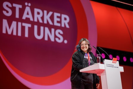 Eine junge Delegierte mit bunten Haarsträhnen steht lächelnd am Rednerpult des DGB-Bundeskongresses. Im Hintergrund ist der Slogan „STÄRKER MIT UNS." zu sehen.
