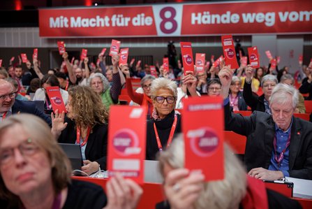 Delegierte des DGB-Bundeskongresses heben rote Stimmkarten während einer Abstimmung im Plenum hoch. Im Hintergrund ist das Motto „Mit Macht für die 8 - Hände weg vom" auf einer großen Leinwand zu sehen.