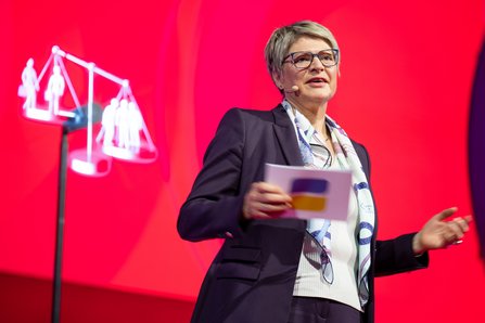 Gundula Roßbach, Präsidentin der Deutschen Rentenversicherung Bund, mit blonder Kurzhaarfrisur, Brille und gemustertem Schal gestikuliert beim Sprechen, im Hintergrund eine rote Leinwand mit Waage-Symbol.