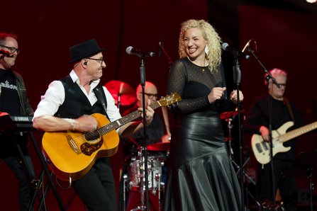 Ein Mann mit Hut und Gitarre und eine Frau mit blonden Locken stehen lachend auf der Bühne vor Mikrofonen, im Hintergrund weitere Bandmitglieder und rote Bühnenbeleuchtung.