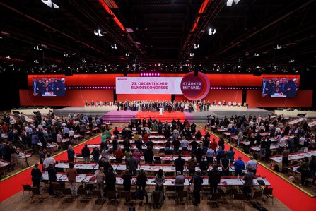 Totale des Kongresssaals: Hunderte Delegierte stehen an ihren Tischen, auf der großen Bühne sind Personen versammelt. Über der Bühne prangt der Schriftzug „23. Ordentlicher Bundeskongress" und „Stärker mit uns." Drei Leinwände zeigen Live-Bilder, rotes Licht taucht die Halle.