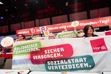 Delegierte in den hinteren Reihen halten ein großes Banner hoch. Darauf steht in bunten Balken: "Solidarisch finanzieren. Sicher versorgen. Sozialstaat verteidigen." sowie das ver.di-Logo. Im Hintergrund das Kongressbanner "Hände weg vom 8-Stunden-Tag!"
