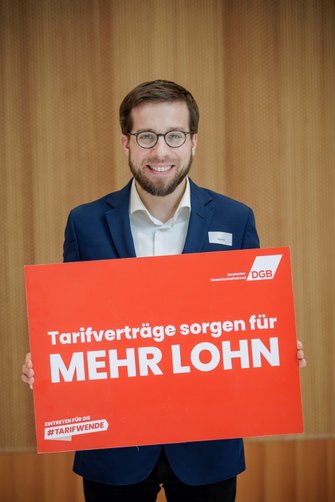 Foto von Victor Perli, ehemaliger Bundestagsabgeordneter für die Linke, der ein Plakat vom DGB in der Hand hält mit der Aufschrift "Tariflöhne sorgen für mehr Lohn".