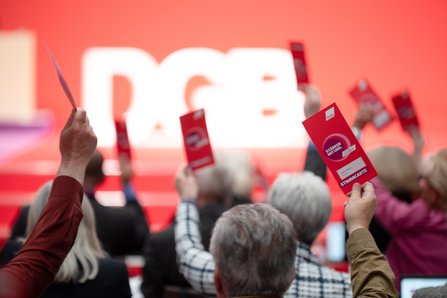 Aus der Perspektive der Delegierten: Mehrere Hände halten rote Abstimmungskarten in die Höhe. Im Hintergrund ist verschwommen das DGB-Logo vor einer Leinwand zu sehen.