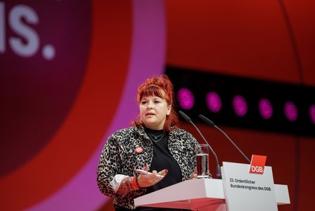 Eine Rednerin mit roten Haaren und Leopardenmuster-Jacke spricht am Pult des DGB-Bundeskongresses. Sie gestikuliert während ihrer Rede.