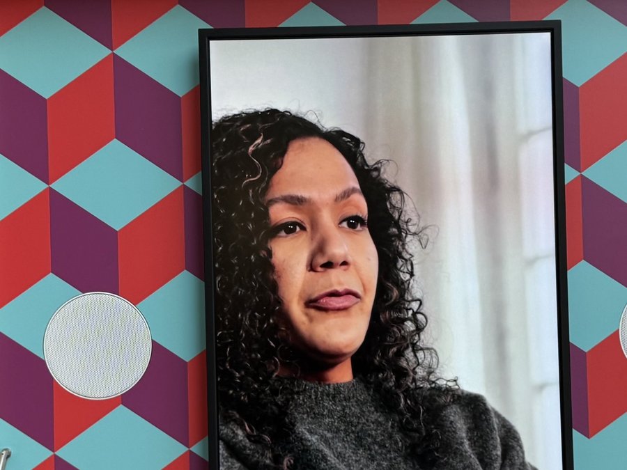 Das Bild zeigt die Ausstellungseinheit "Klingel" der Vielfaltsausstellung. Die Einheit besteht aus einem Holzrahmen mit einem Bildschirm, der von einem geometrischen Muster aus roten und blauen Formen umgeben ist. Die Einheit steht auf einem gelben Sockel, der mit dem DGB-Logo versehen ist.