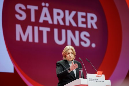Eine Frau mittleren Alters mit blonden Haaren steht am Rednerpult und spricht. Sie trägt einen schwarzen Blazer über einem grauen Rollkragenpullover. Im Hintergrund der Schriftzug "Stärker mit uns".