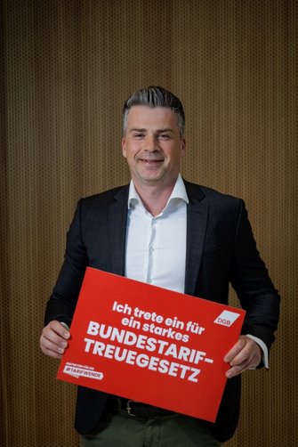 Foto von Robby Riedel, der ein rotes Schild in der Hand hält mit der Aufschrift "Ich trete ein für ein starkes Bundestariftreuegesetz"