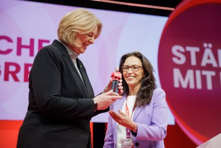 Zwei Frauen auf der Bühne. Eine Frau in schwarzem Blazer überreicht einer lächelnden Frau im hellvioletten Blazer eine Miniatur-Figur. Im Hintergrund ist der Schriftzug "Stärker mit uns" zu sehen.