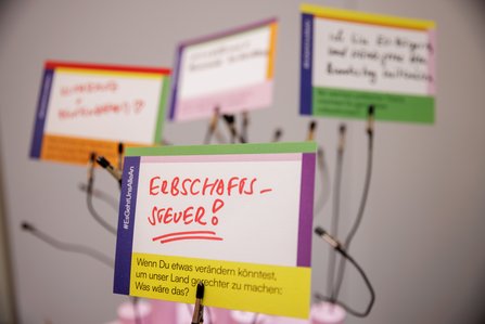 Bunte Moderationskarten mit handgeschriebenen Fragen an einer Pinnwand. Im Vordergrund eine Karte mit der Aufschrift „Erbschaftssteuer?" in roter Schrift, darunter der Text „Wenn Du etwas verändern könntest, um unser Land gerechter zu machen: Was wäre das?" und der Hashtag #EGBnurSAGEN.