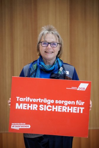 Foto von Hannelore Buls, ehemalige Vorsitzende des deutschen Frauenrats, die ein rotes Plakat vom DGB in den Händen hält mit der Aufschrift "Tarifverträge sorgen für mehr Sicherheit".