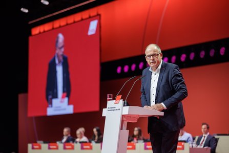 Stefan Körzell mit Brille im Anzug steht am Rednerpult und spricht, im Hintergrund ist eine große rote Leinwand und er auf einer Projektionsfläche zu sehen.