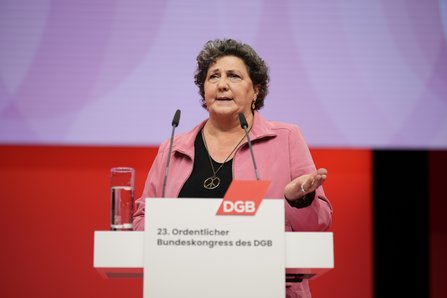 Eine Frau mit lockigem braunem Haar und rosafarbener Jacke steht am Rednerpult und gestikuliert mit der rechten Hand. Vor ihr stehen zwei Mikrofone, im Hintergrund ist eine rot-violette Leinwand zu sehen.