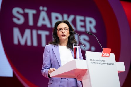 ine Frau mit dunklen, schulterlangen Haaren und Brille spricht am Rednerpult. Sie trägt einen hellvioletten Blazer über einem weißen Oberteil. Im Hintergrund der Schriftzug "Stärker mit uns".