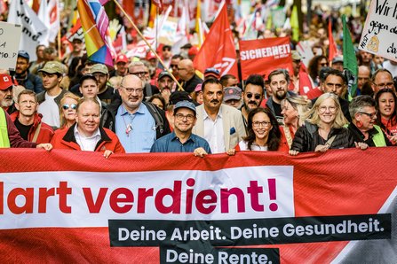 DGB-Vorsitzende Yasmin Fahimi steht mit weiteren Gewerkschaftsvertreter*innen hinter dem Transparent „Hart verdient!“; dahinter demonstriert eine dicht gedrängte Menschenmenge.