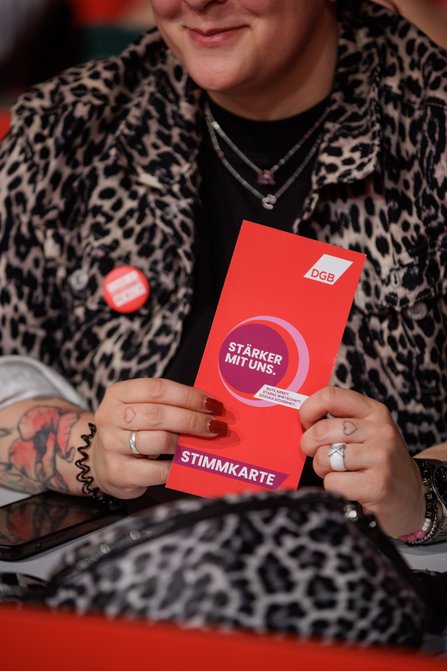 Nahaufnahme einer Person mit Leopardenmuster-Jacke, mehreren Halsketten und Tattoos, die eine rote DGB-Stimmkarte mit dem Slogan "STÄRKER MIT UNS" in den Händen hält.
