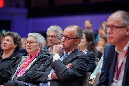 Mehrere Personen sitzen konzentriert im Publikum. Im Vordergrund ein Mann im Anzug mit der Hand am Kinn, der nachdenklich nach vorne schaut. Neben ihm Frauen mit pinkem Schal und Jacke.