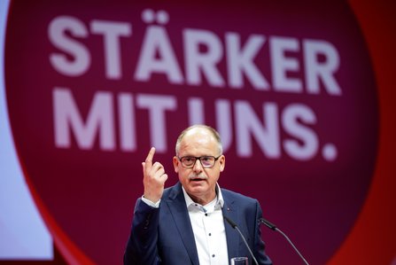 Ein Mann mit Brille und grauem Haar steht am Rednerpult und hebt den rechten Zeigefinger. Hinter ihm ist auf einer großen Leinwand der Schriftzug "STÄRKER MIT UNS." in einem roten Kreis zu sehen.