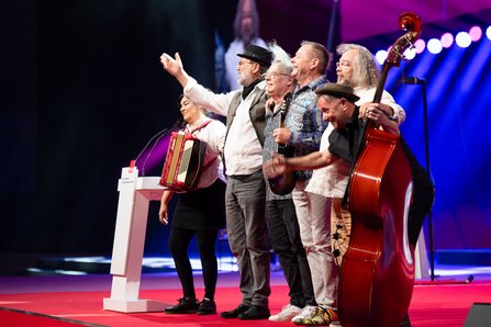 Eine Musikgruppe mit sechs Personen steht auf der Bühne, darunter eine Frau mit Akkordeon und Musiker mit Kontrabass, sie winken und lachen in Richtung Publikum, bunte Bühnenbeleuchtung im Hintergrund.