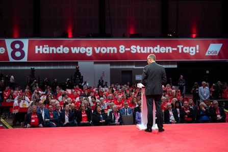 Rückansicht eines Redners im dunklen Anzug am Pult. Er blickt in die mit Delegierten gefüllte Kongresshalle. Im Hintergrund ein großes rotes Banner: "Hände weg vom 8-Stunden-Tag!"