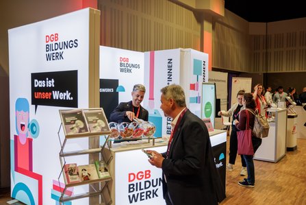 Zwei Männer stehen sich am Stand des DGB Bildungswerks gegenüber. Links ein Mann mit rotem Lanyard, rechts ein Mann mit violettem Lanyard. Im Hintergrund bunte Stellwände mit der Aufschrift „Das ist unser Werk."