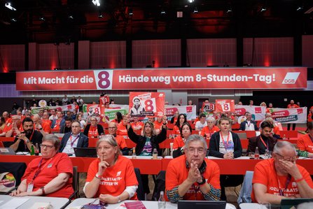 Kongresshalle beim 23. Ordentlichen DGB-Bundeskongress. Im Vordergrund sitzen Delegierte an roten Tischen, viele tragen T-Shirts mit der Aufschrift "Sozialstaat verteidigen". Im Hintergrund ist ein großes Banner zu sehen: "Mit Macht für die 8 – Hände weg vom 8-Stunden-Tag!" Delegierte halten Schilder und Plakate hoch.