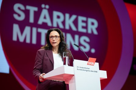 Eine Frau mit langen dunklen Haaren und Brille in violettem Hosenanzug steht am Rednerpult und hält Papiere in der Hand. Hinter ihr ist auf einer großen Leinwand der Schriftzug „STÄRKER MIT UNS." in einem roten Kreis zu sehen.