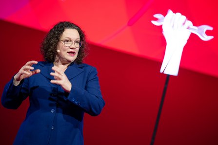 Andrea Nahles gestikuliert lebhaft vor einem roten Hintergrund mit einer weißen Faust als Hologramm, im blauen Kleid und mit Brille.
