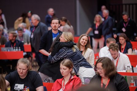 Zwei Personen umarmen sich in den Sitzreihen eines Kongressraums, im Hintergrund sind Delegierte zu sehen, die sitzen und aufmerksam wirken; die Szene zeigt Nähe und Wiedersehen.