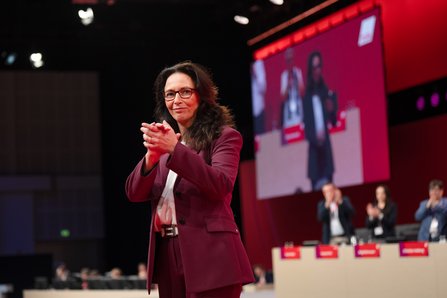 Eine Frau mit langen dunklen Haaren und Brille in violettem Hosenanzug steh vor dem Präsidium. Im Hintergrund sind auf einer Leinwand weitere Personen zu sehen, die applaudieren.v
