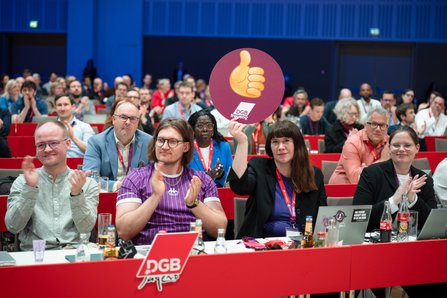Mehrere Delegierte sitzen an roten Tischen im Kongresssaal. Eine Frau in der Mitte hält ein rundes Schild der DGB-Frauen mit einem Daumen-hoch-Symbol hoch. Im Hintergrund applaudieren weitere Delegierte.
